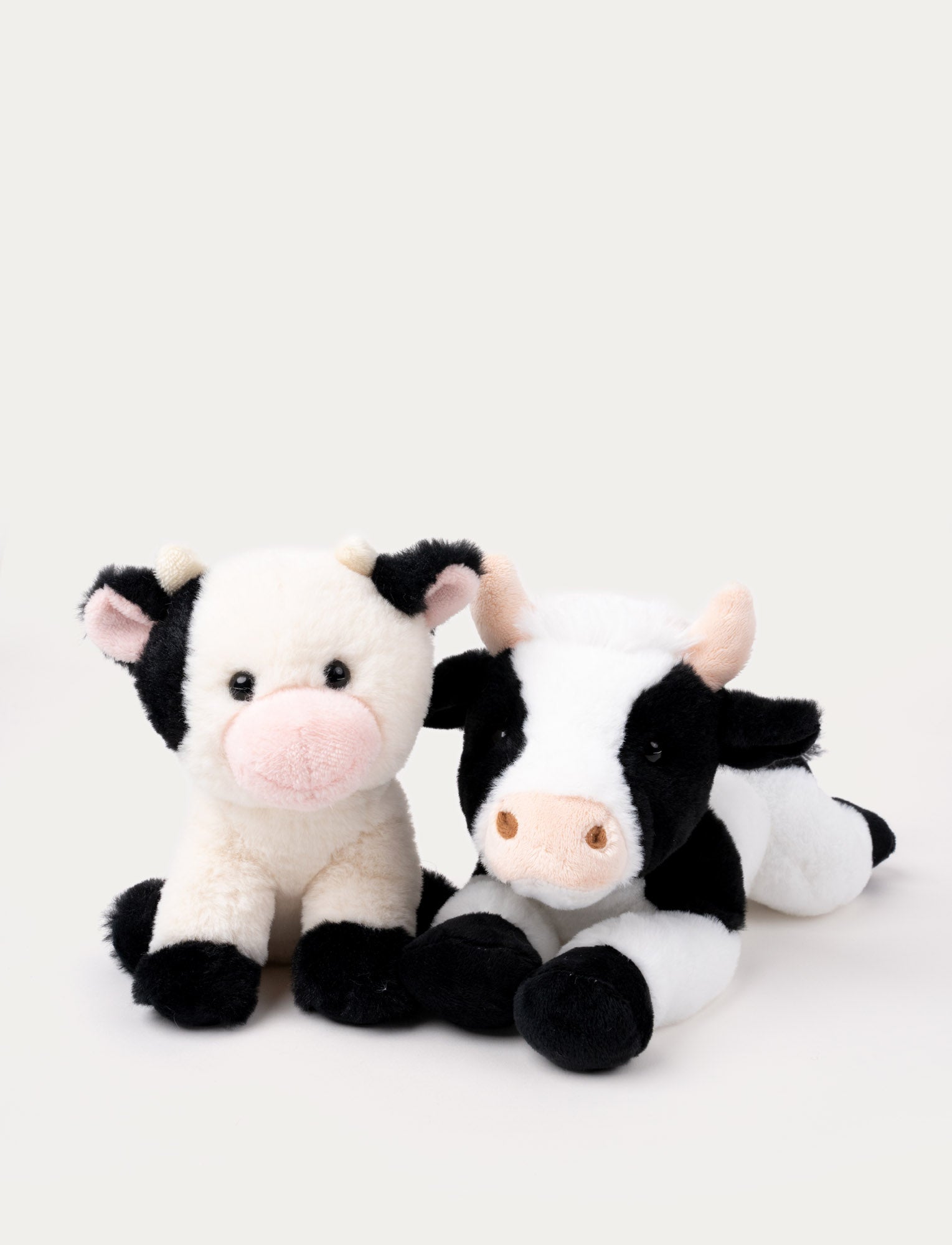 Image of Two plush cow toys, one sitting with a pink nose and the other lying down with a black-and-white pattern.