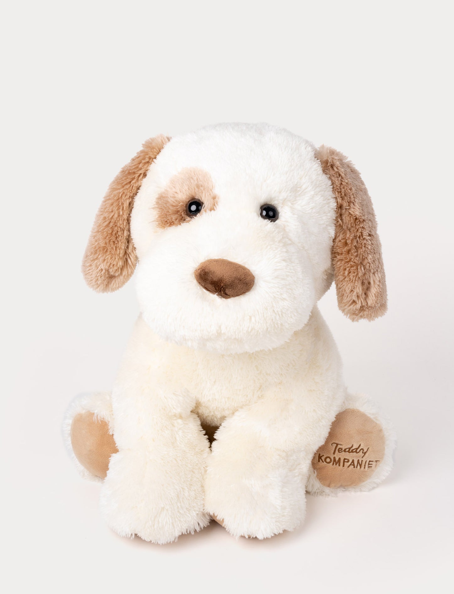 Soft plush dog with cream-colored fur, brown floppy ears, a brown patch over one eye, and "Teddy Kompaniet" on one paw.