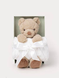 Gift set with a beige plush teddy bear holding a soft white blanket tied with a satin ribbon, presented in a green box.